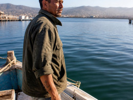A weathered Mexican man stands on a fishing boat at harbor with Ensenada coast behind, his face serious and eyes squinting at the sea.