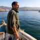 A weathered Mexican man stands on a fishing boat at harbor with Ensenada coast behind, his face serious and eyes squinting at the sea.