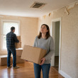 Woman looking up at a crooked ceiling vent in an empty living room while holding a box, man in background carrying boxes.
