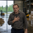 Middle-aged man in warehouse pressing hand to chest, looking tense.