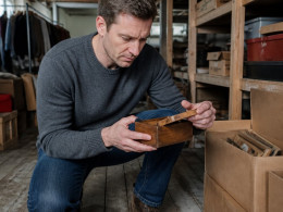Man crouching in a cluttered basement holding a small cigar box, looking intently inside.