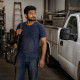 South Asian man in his 30s with a tool bag over his shoulder in a garage, grease on his forearm.
