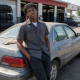 Evan leaning on a sedan outside a tire shop, grease on his hands, work shirt.